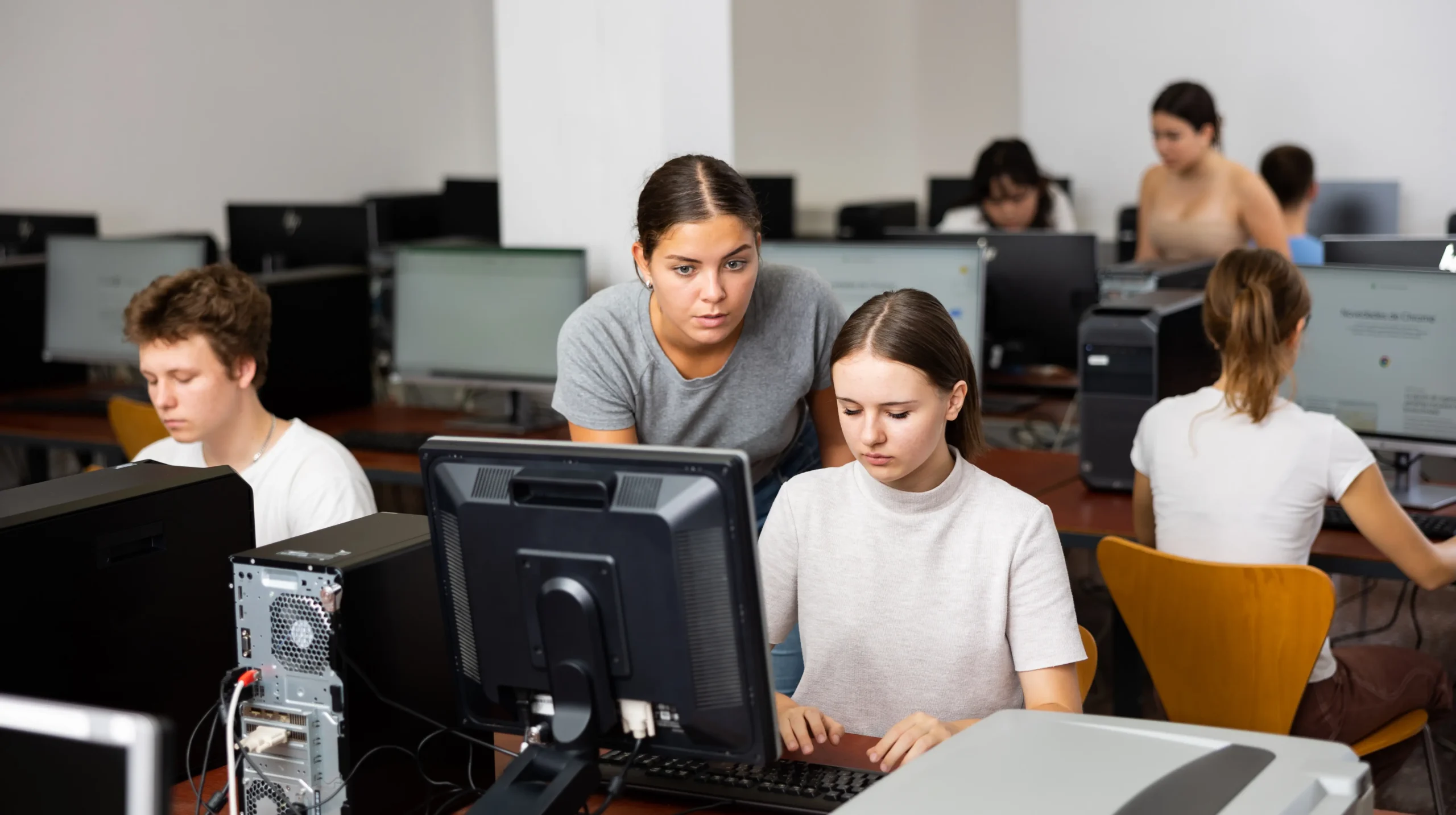 Students working on desktop computers in a computer lab while receiving guidance from an instructor.